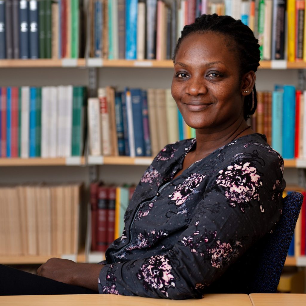 Betty Nannyonga sitter i ett rum fyllt med bokhyllor. Hon ler och tittar in i kameran. Hon har en grå blus med rosa blommor.