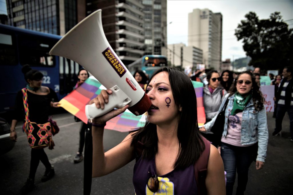 En kvinna går i ett demonstrationståg mot våld mot kvinnor i Quito, Ecuador. Hon håller upp en megafon mot ansiktet. I bakgrunden syns fler demonstranter.