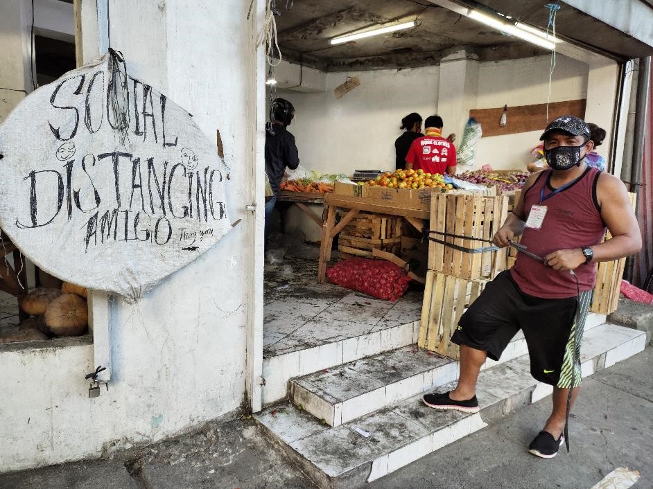 En sommarklädd man med ansiktsmask står vid en marknad i staden Manila på Filippinerna. På väggen bredvid honom sitter en skylt uppsatt med texten "Social distancing, amigo"
