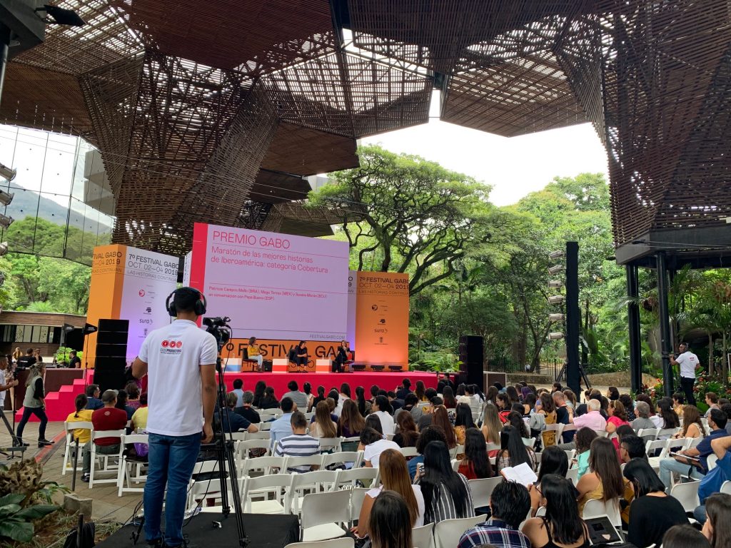 En kameraman står på ett podium i förgrunden och riktar sin kamera mot en scen i bakgrunden. Framför scenen sitter en publik på vita stolar. På scenen står det textat "Festival Gabo".