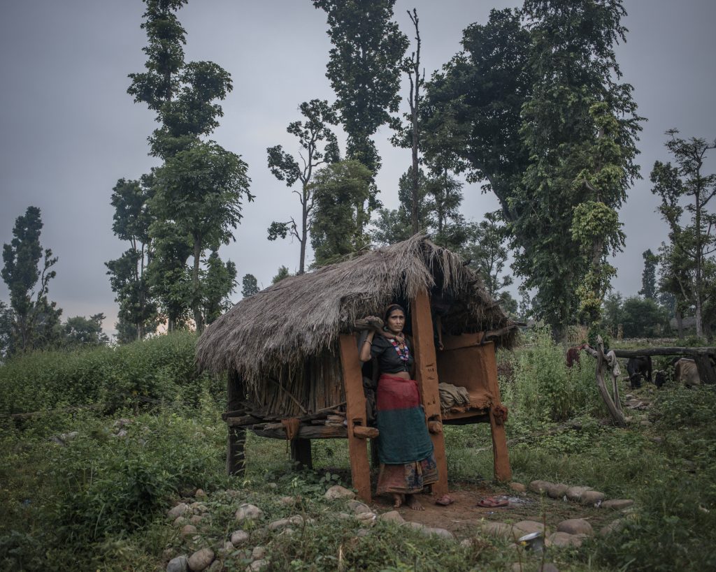 Chandra, en kvinna, står på ett fält, framför en så kallad menshydda. Det är en enkel hydda i trä och med halmtak. Bakom henne syns buskar och träd.