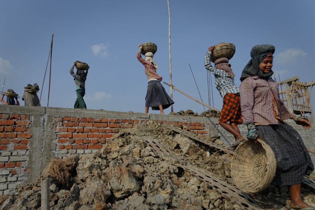 Byggarbetare i Myanmar bär byggmaterial i flätade korgar. De går på rad över en hög av bråte och en tegelmur.