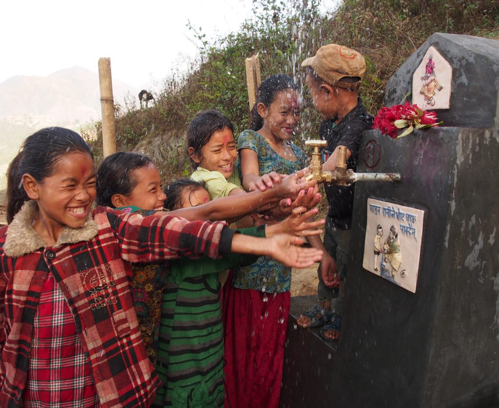 Fem barn leker vid en nyinstallerad vattenkran. I bakgrunden syns berg och växtlighet.