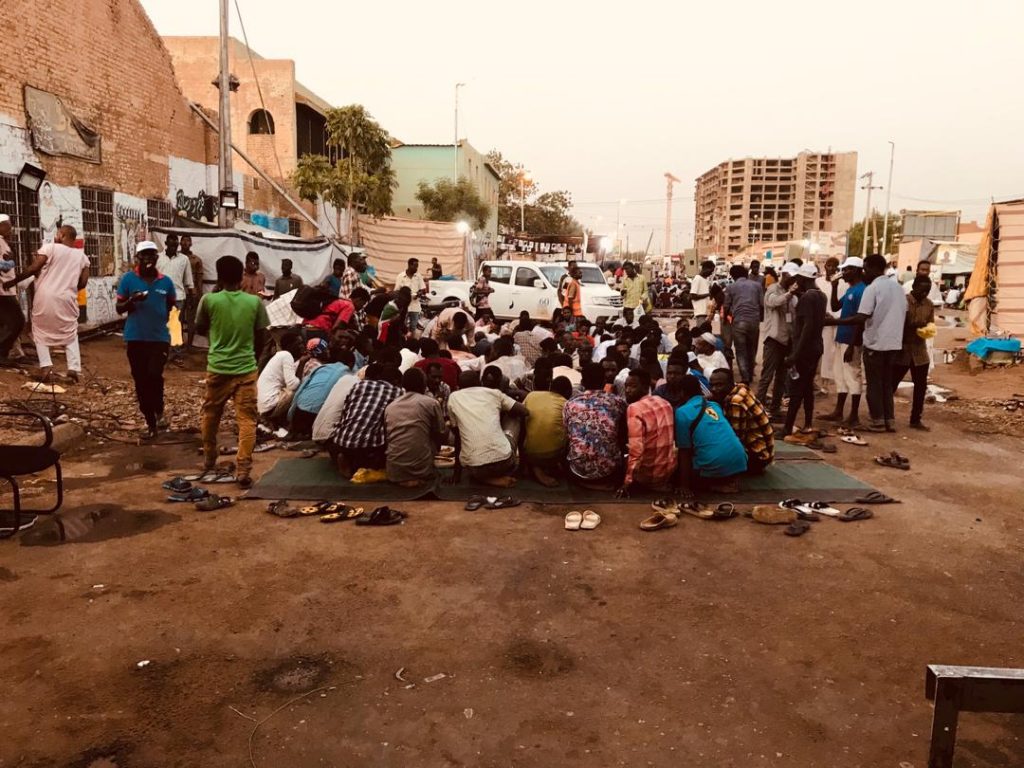 Drygt femtio protestanter sitter på filtar på marken. Runt om dem står människor. I bakgrunden syns byggnader, träd och en bil.