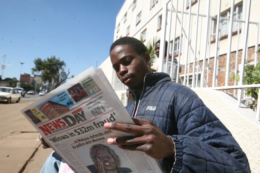 En man sitter på en bänk och läser tidningen 'NewsDay'. Bakom honom syns byggnader och bilar.