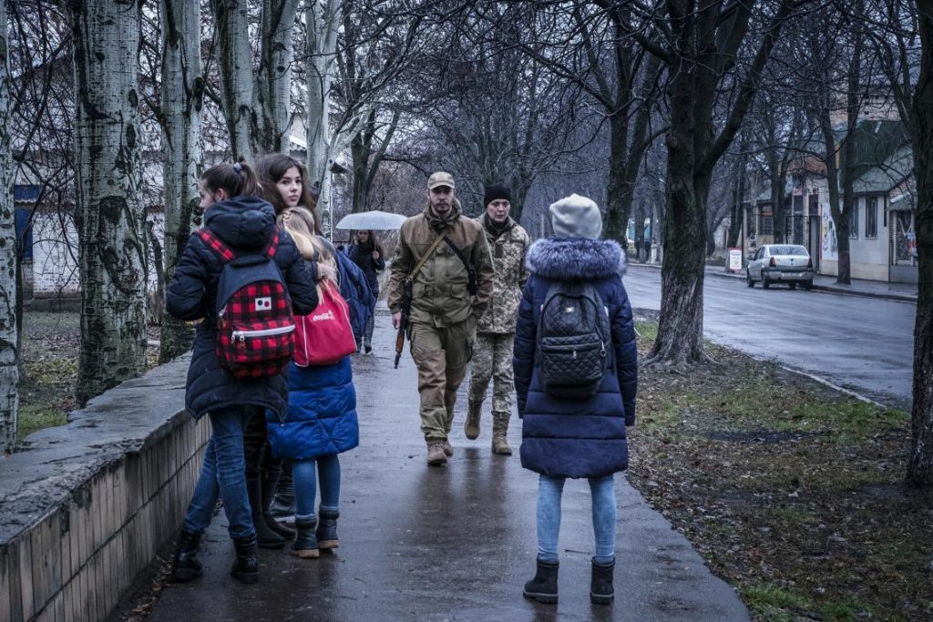 Skolbarn och militärer passerar varandra på en trottoar. I bakgrunden syns hus och träd.