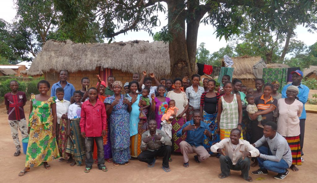 Ett 30-tal män, kvinnor och barn står eller sitter framför ett tegelhus med halmtak för att bli fotograferade. Kvinnorna har färgglada klänningar eller kjolar. De tillhör en Dimitraklubb i DRC Kongo.