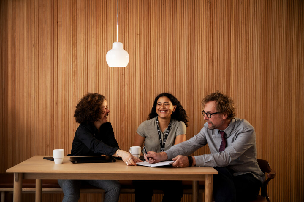 Två kvinnor och en man sitter vid ett bord framför en träpanel.