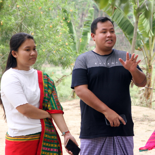 En man står framför en grupp och pratar i en lantlig miljö. Bredvid honom står även två kvinnor i traditionella kläder.