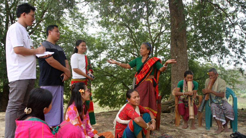 Några personer diskuterar i grupp utomhus. En kvinna viftar med händerna medan hon pratar. Några sitter på marken och andra står upp.