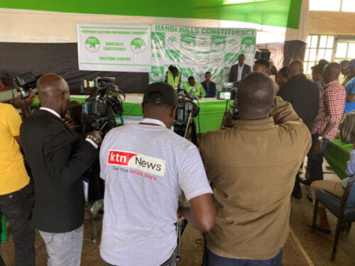 En presskonferens där fotografen har tagit bilden bakom ryggen på 3 kameramän, mot en scen där det står 4 personer.