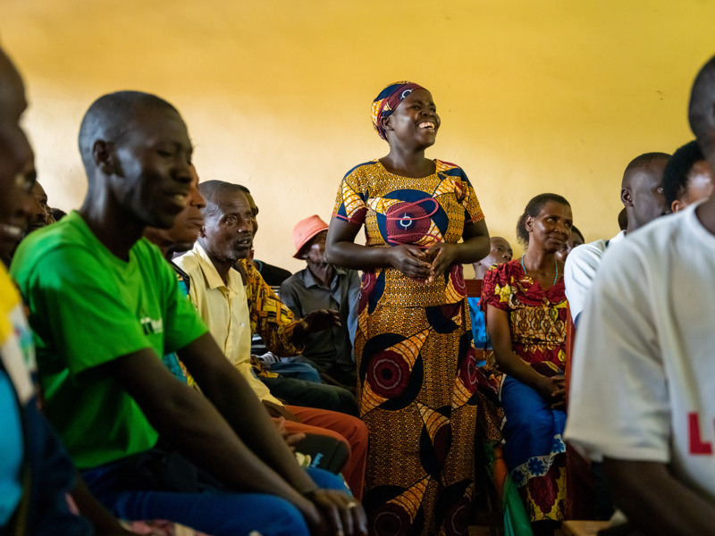 En kvinna står upp från sin stol i en stor folksamling inomhus. Hon ler stort.