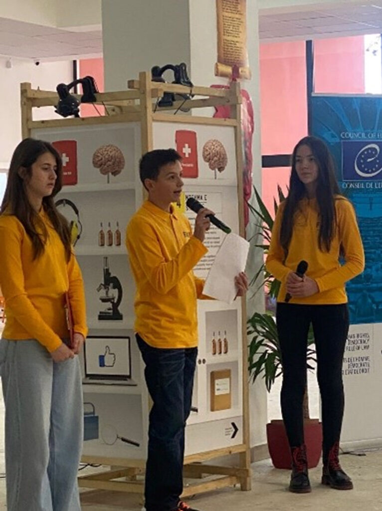 Tre ungdomar i matchande gula tröjor står vid en monter och håller i mikrofoner. Pojken i mitten pratar i sin och läser på ett papper.