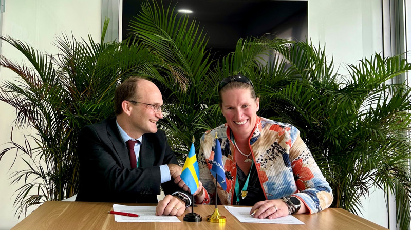 En man och en kvinna sitter vid ett bord framför gröna växter och skakar hand. De ler stort.