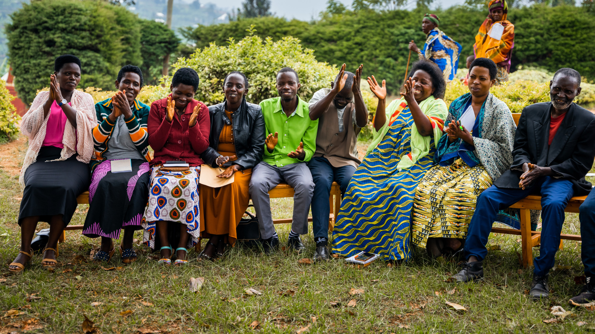 En grupp människor sitter på träbänkar på en gräsmatta och klappar händerna. De ser glada ut.