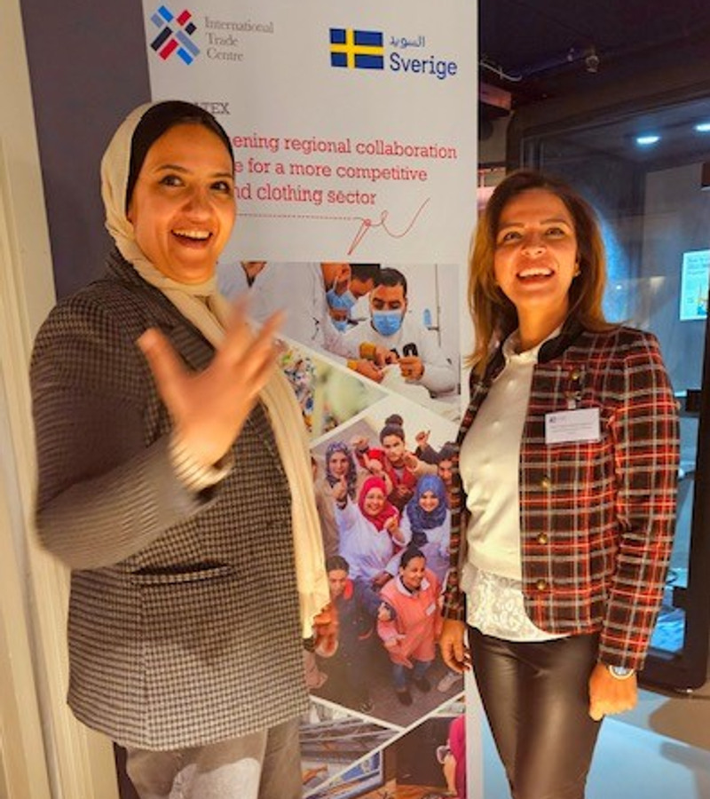 Två kvinnor står framför en banderoll för hållbar klädindustri och ler stort mot kameran.