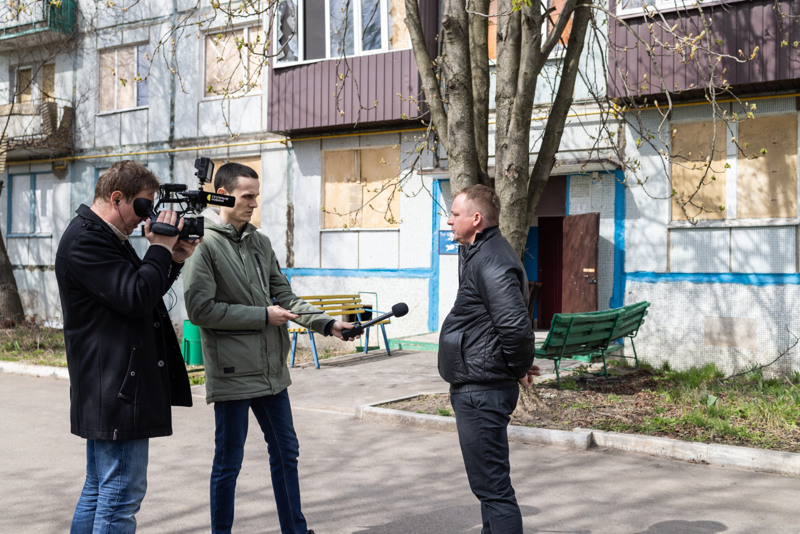 Två journalister filmar och håller fram en mikrofon till en man. I bakgrunden ett flerfamiljshus.