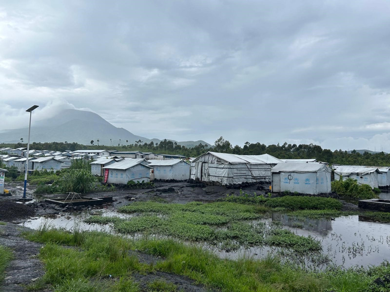 Ett stort läger bestående av vita tält märkta med UNHCR. I förgrunden gräs och pölar, i bakgrunden berg. Det regnar.