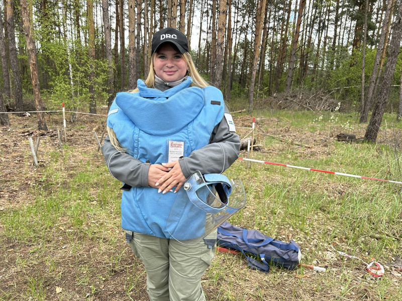 Porträtt taget framför en tallskog på en kvinna som bär en stor ljusblå skyddsväst och svart keps. Hon håller i ett skyddsvisir.