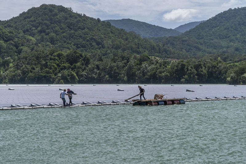 Ett fält med flytande solpaneler på en stor sjö med gröna berg i bakgrunden. Tre personer arbetar på solpanelerna.