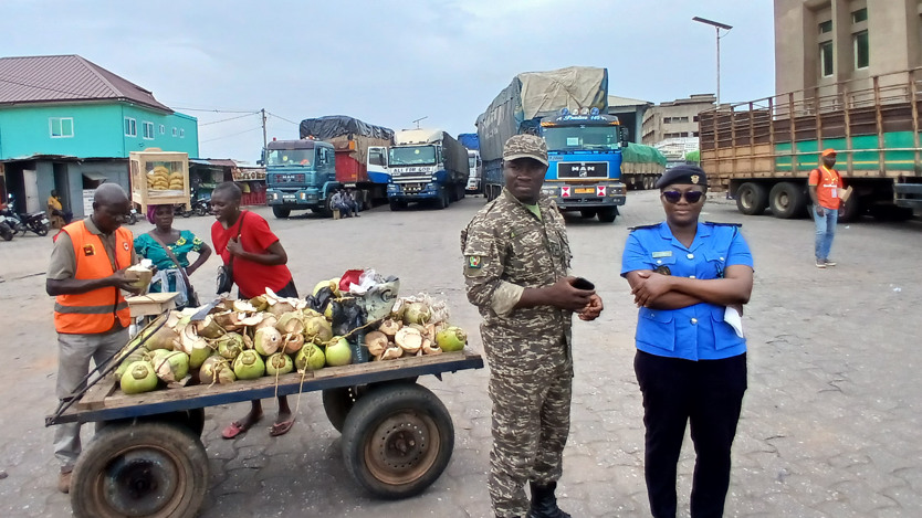 Ett torg med en vagn med grönsaker på och flera personer som står utspridda, varav några har uniform. I bakgrunden står flera lastbilar.
