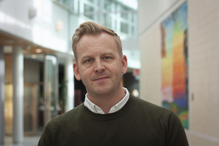 Närbild på man i grön stickad tröja med vit skjortkrage som sticker upp.