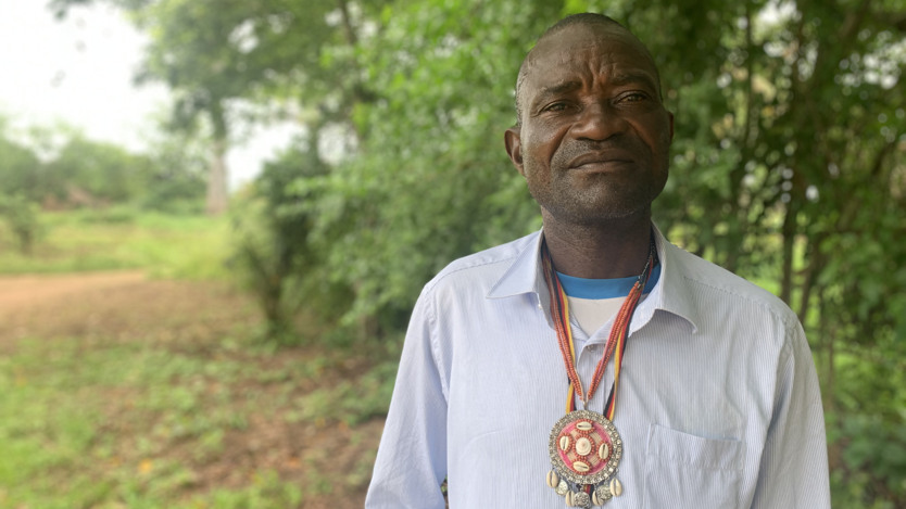 En man som står framför en skog. Rund halsen har han en medaljong.