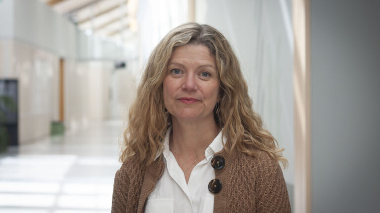 Porträtt på kvinna i långt vågigt hår, vit skjorta och brun stickad kofta.