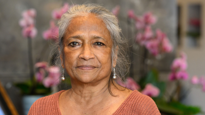 Kvinna med grått hår tittar rakt in i kameran och har en mörkt aprikos tröja på sig.