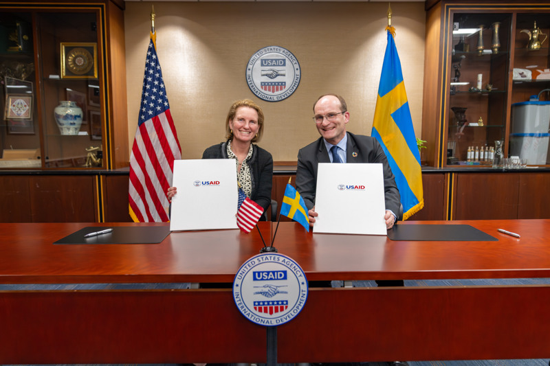 En man och en kvinna sitter vid ett skrivbord med en svensk och en amerikansk flagga vid sina sidor.