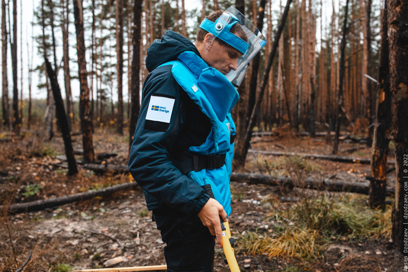 En man i blå skyddsväst och genomskinligt skyddsvisir står i en skog en mulen dag. Han håller i en orange stav och tittar ner i marken.