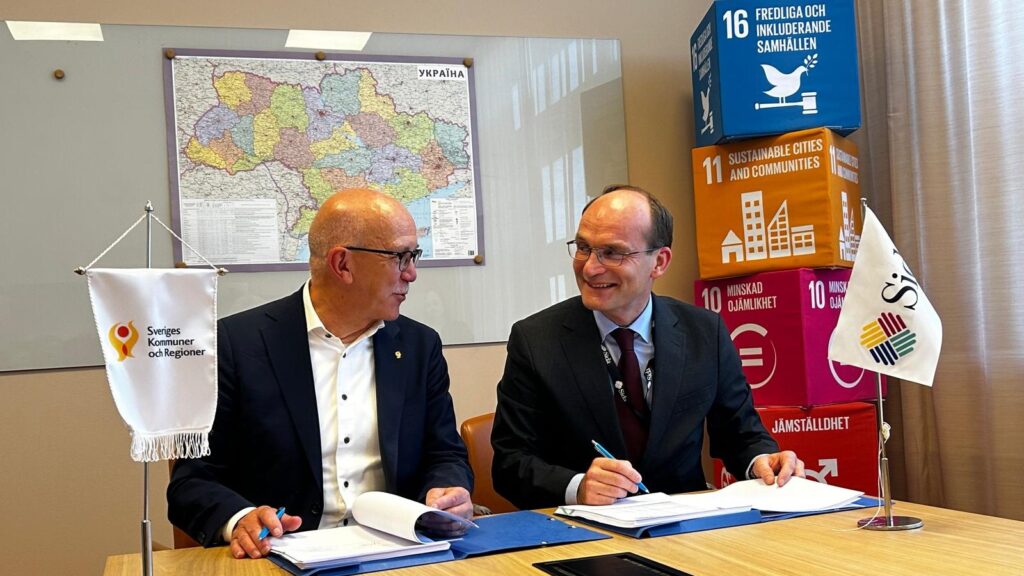 Två män sitter vid ett skrivbord och tittar mot varandra och ler.