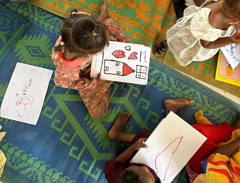 Barn sitter på en matta och håller i teckningar som de har ritat.