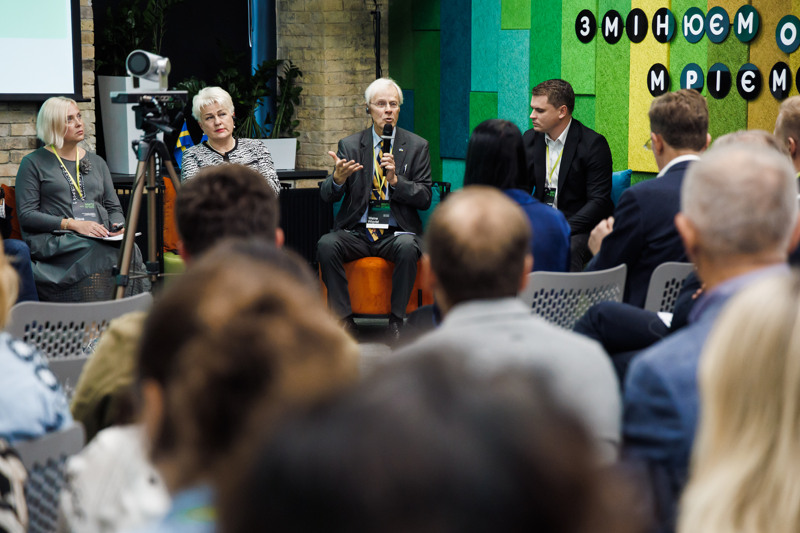 Fyra personer sitter på en låg scen framför en publik. En man pratar i en mikrofon.