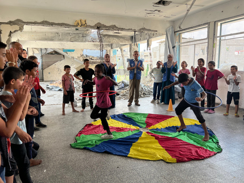Två barn rockar med rockringar på en färgglad matta. Andra barn står vid sidan om och tittar och applåderar.