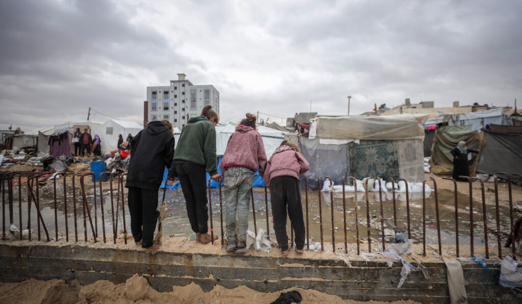 Fyra barn står vid ett räcke och tittar ut över tält  och hus som skadats.