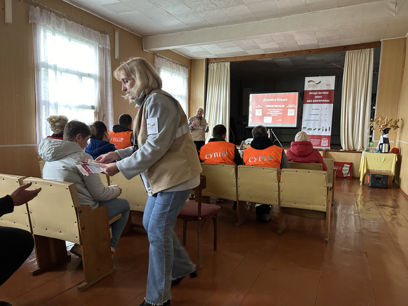 En mötessal med personer sittande på stolar som lyssnar på en föreläsare längst fram. En kvinna går runt och delar ut en broschyr.