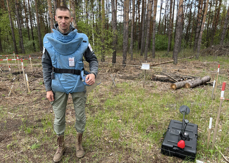 Porträtt taget framför en tallskog på en man som bär en stor ljusblå skyddsväst. Han håller i ett skyddsvisir.