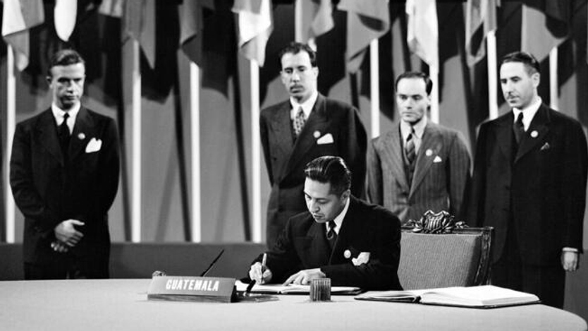 En grupp män som står runt ett bord och skriver under papper. I bakgrunden står flaggor uppställda.