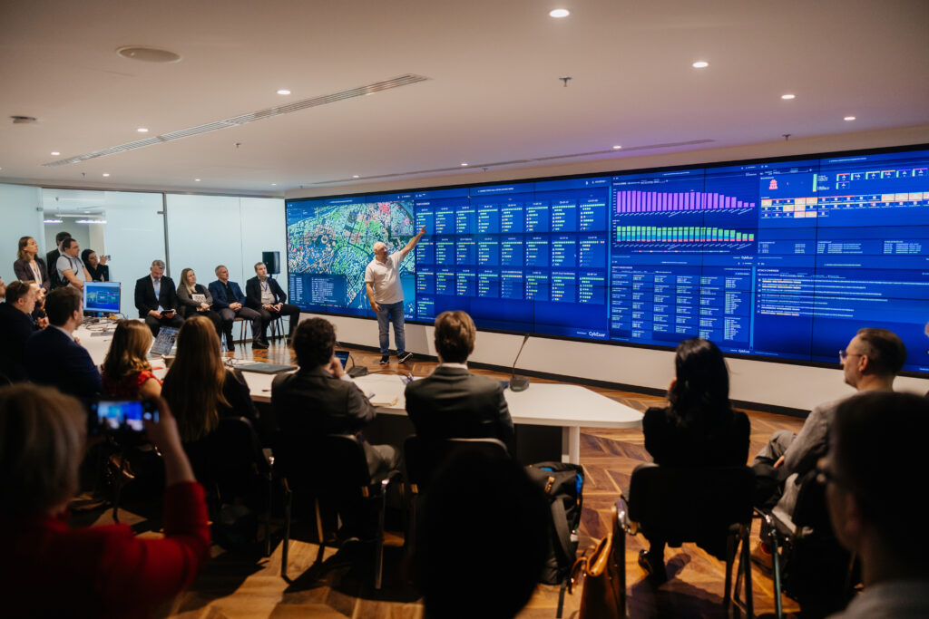 En stor grupp människor som sitter framför en skärm som tar upp en hel vägg. En man står och presenterar något på skärmen.