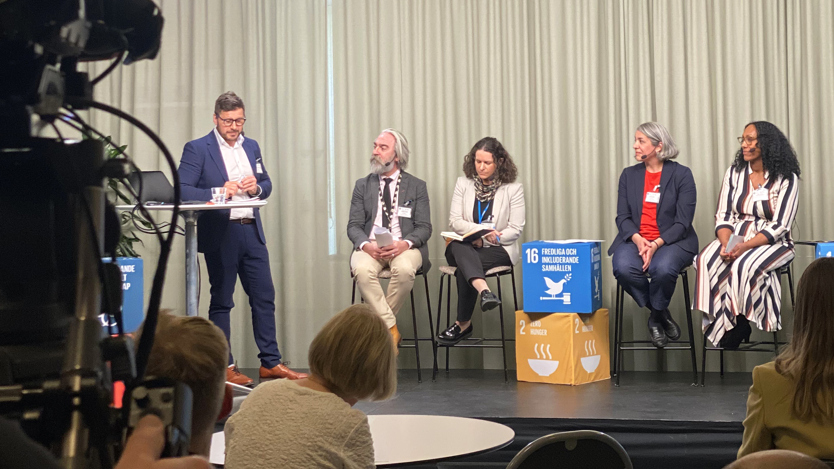 En grupp människor som sitter på en scen. Framför scenen sitter några och lyssnar.