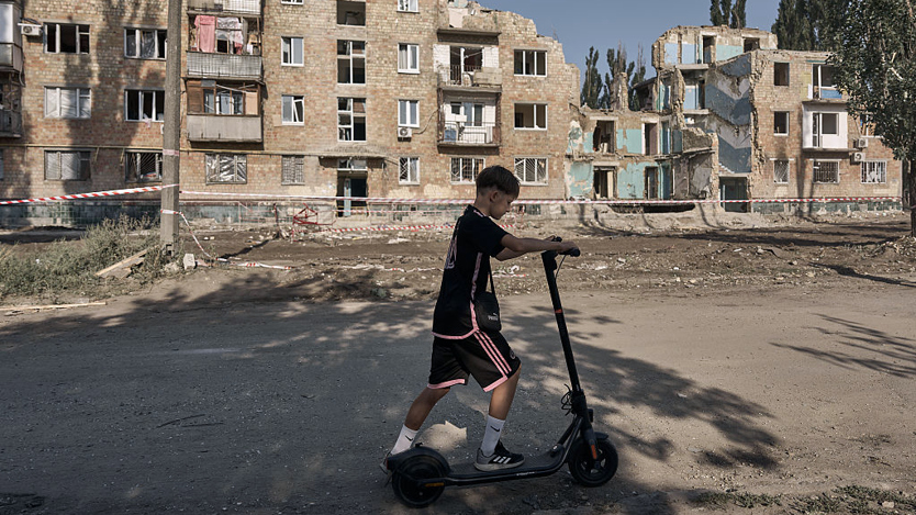 Ett barn på sparkcykel framför sönderbombade hus.