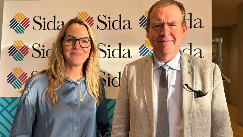 Två personer, en kvinna med långt blont hår i blå skjorta och en man med kort ljust hår och kavaj,  står framför en vepa där det står Sida.