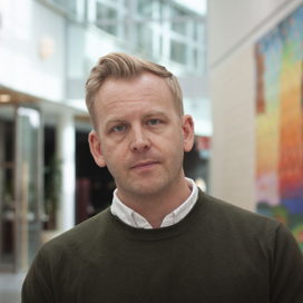 Närbild på man i grön stickad tröja med vit skjortkrage som sticker upp.