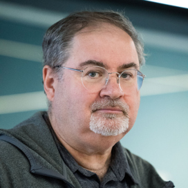 Halvfigursporträtt på man i grått skägg, glasögon och grå luvtröja.