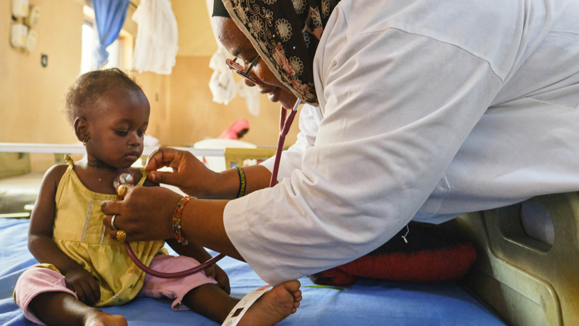 En kvinna i vit rock lyssnar på ett litet barns hjärta med stetoskop. Barnet sitter i en sjukhussäng.