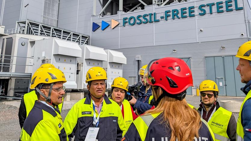 En grupp människor står i varselkläder och hjälm framför en fabrik med texten Fossil Free Steel.